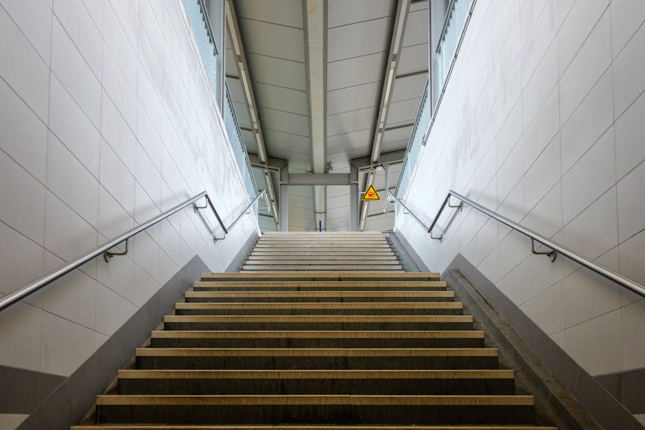 A wide staircase inside a commercial or public building, leading up to an exit with a glass wall at the top. The stairs are made of dark wood or laminate with light-colored edging strips, and there are stainless steel handrails on both sides mounted on smooth, tiled walls. The staircase is well-lit with natural light coming through the glass wall, and a yellow warning sign is visible near the top, indicating caution for movement or obstacles. The environment appears to be part of a home relocation or moving process managed by Removal Companies Stockwell, with the staircase facilitating the transport of furniture, boxes, or appliances during the packing and loading phases of a house removal. The setting suggests readiness for furniture transport through the building’s stairs, with attention to safety and organization for efficient moving logistics.