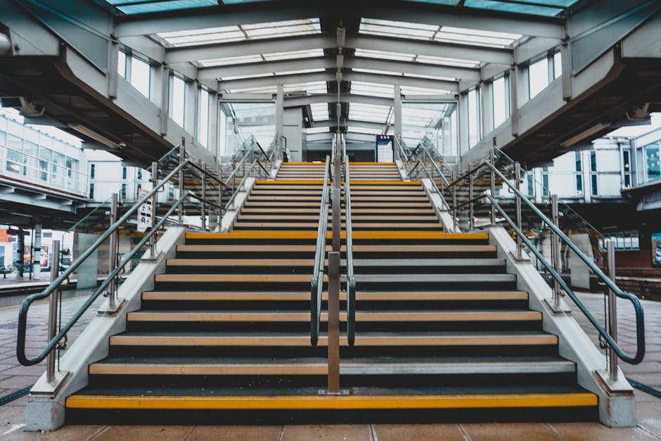 Exterior view of a modern train station stairway with detailed wooden steps featuring black anti-slip strips and yellow safety lines along the edges. The staircase has sleek metal handrails on both sides and a central handrail running down the middle, leading up to a covered glass and metal structure with natural daylight filtering through. The scene showcases a spacious, well-lit environment suitable for home relocation activities involving the transport of furniture, boxes, and packing materials, with the station serving as a potential point for moving logistics. The image emphasizes the technical aspects of navigating narrow staircases during a house removal, highlighting the importance of careful packing, strategic lifting, and efficient furniture transport between the station and nearby transport vans, which might be parked outside, ready for loading. This visual aligns with professional removals services like those offered by Removal Companies Stockwell, particularly focusing on challenges posed by stairs during a house move in a busy urban setting.
