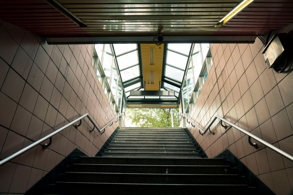 A wide staircase inside a commercial or public building, leading up to an exit with a glass wall at the top. The stairs are made of dark wood or laminate with light-colored edging strips, and there are stainless steel handrails on both sides mounted on smooth, tiled walls. The staircase is well-lit with natural light coming through the glass wall, and a yellow warning sign is visible near the top, indicating caution for movement or obstacles. The environment appears to be part of a home relocation or moving process managed by Removal Companies Stockwell, with the staircase facilitating the transport of furniture, boxes, or appliances during the packing and loading phases of a house removal. The setting suggests readiness for furniture transport through the building’s stairs, with attention to safety and organization for efficient moving logistics.